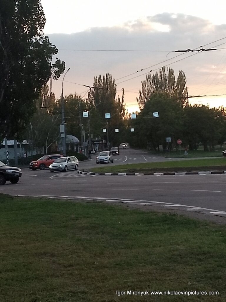 Когда солнце уже село за горизонт, настают сумерки. В Николаеве, на фото видно городской трафик и зелёные газоны и деревья.