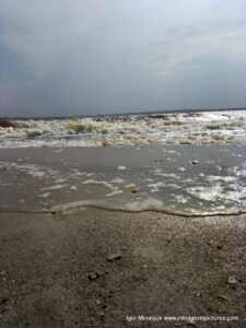 На фото бущующяя вода, уходящая волна, мокрый песок и затянутое облаками небо.