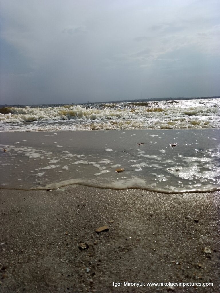 На фото бущующяя вода, уходящая волна, мокрый песок и затянутое облаками небо.