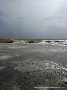 На фото бущующяя вода, уходящая волна, мокрый песок и затянутое облаками небо.