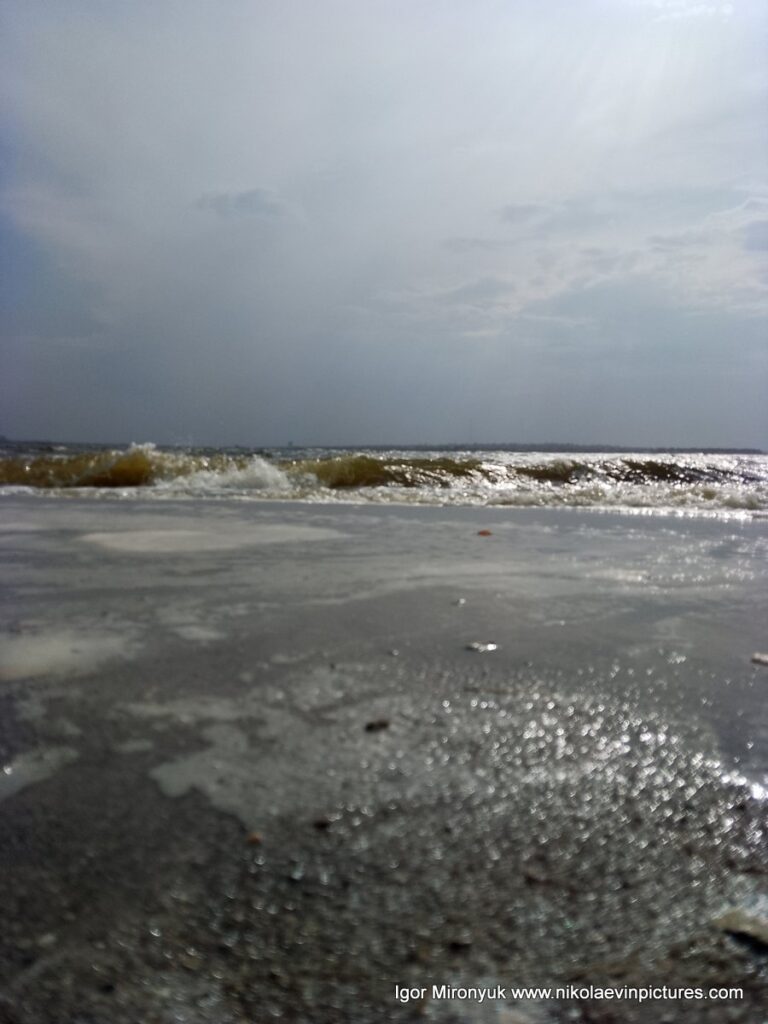 На фото бущующяя вода, уходящая волна, мокрый песок и затянутое облаками небо.